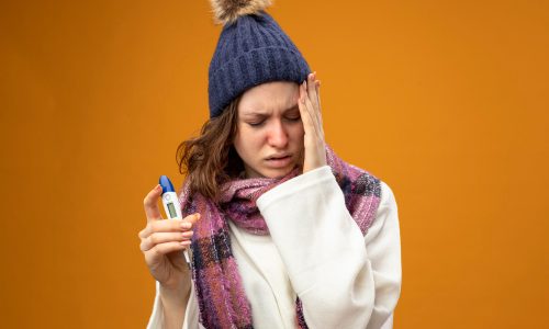 unpleased-young-ill-girl-with-closed-eyes-wearing-white-robe-winter-hat-with-scarf-holding-thermometer-putting-hand-cheek-isolated-orange